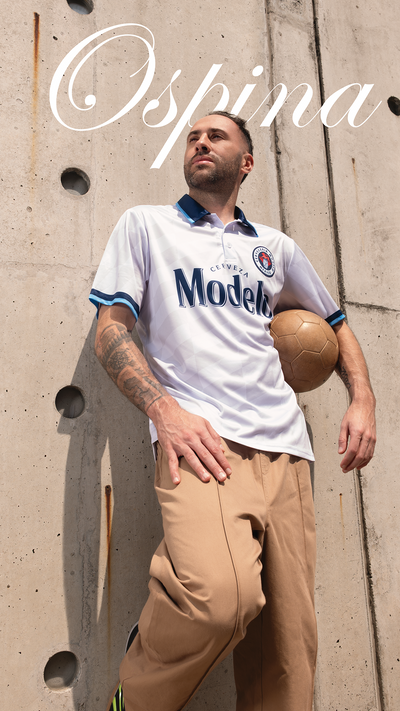 David Ospina wearing a white soccer jersey with 'Modelo' branding against a concrete wall.