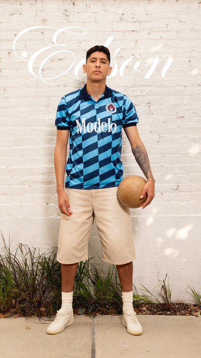 Edson Álvarez wearing a blue checkered soccer jersey with 'Modelo' branding, standing against a white brick wall.